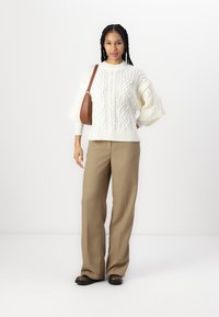 Maglione bianco a cuiacchi con maniche ampie, abbinato a pantaloni beige a vita alta e una borsa a spalla marrone. Scarpe scure completano l'outfit.