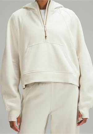 Person iført cremefarvet cropped hoodie med halv lynlås og frontlomme, kombineret med matchende høj talje bukser.