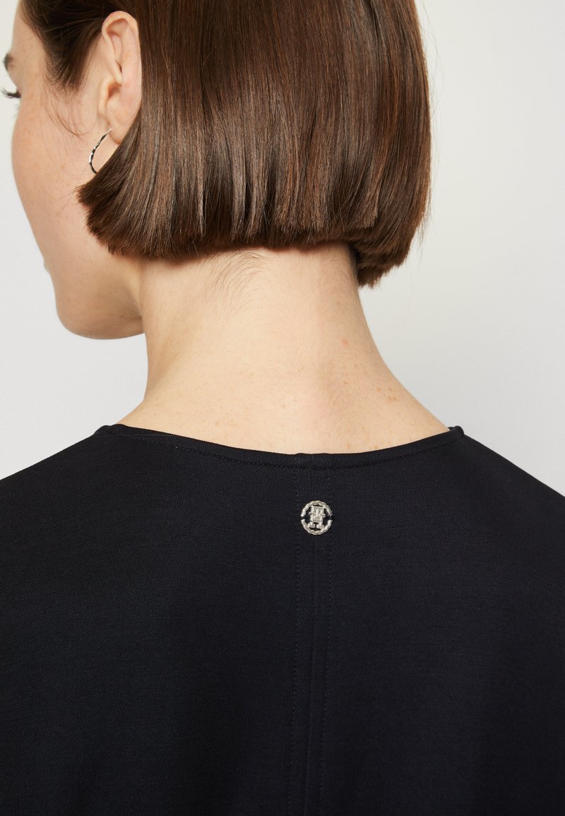 Frau mit kurzem braunem Haar, die ein schwarzes Top trägt, das oben am Rücken, nahe dem Ausschnitt, ein kleines rundes silbernes Emblem hat.