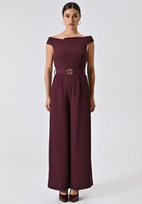 Burgunderfarbener Jumpsuit mit schulterfreiem Design, weitem Bein und taillierter Passform, verziert mit einer dekorativen goldfarbenen Schnalle. Glatte Textur.