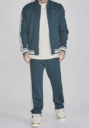Homme portant un survêtement bleu foncé avec des bandes blanches, des baskets blanches et une chemise blanche, debout devant un fond uni.