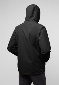 Persona vestida con una chaqueta con capucha negra y las manos en los bolsillos, vista desde atrás contra un fondo gris liso.