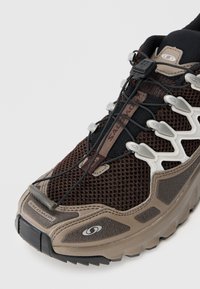 Καφέ και μπεζ ορειβατικό παπούτσι με επάνω από δίχτυ, σύστημα κορδόνι με κωδωνοκρουσία και ανθεκτική σόλα. Διαθέτει έντονη σήμανση και υφή σε διάφορες λεπτομέρειες.