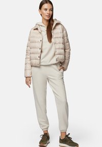 Nabłyszczana beżowa kurtka z przekrojonym fasonem, w połączeniu z jasnobeżowym hoodie i dopasowanymi joggerami. Zielone oliwkowe sneakersy dopełniają wygląd.