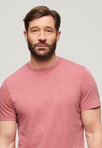 T-shirt a maniche corte di colore rosa sbiadito, realizzato in tessuto texturizzato. Presenta un collo rotondo e una vestibilità casual con dettagli di cucitura in evidenza.
