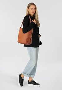 Borsa tote in pelle marrone con finitura testurizzata, caratterizzata da un accento intrecciato su un lato e una tracolla resistente. Il modello indossa un outfit casual.