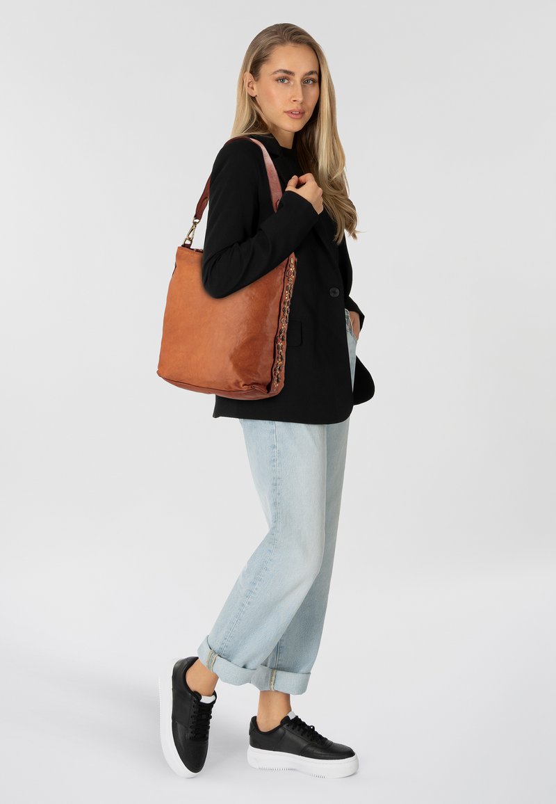 Borsa tote in pelle marrone con finitura testurizzata, caratterizzata da un accento intrecciato su un lato e una tracolla resistente. Il modello indossa un outfit casual.