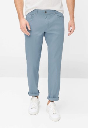 Mand iført lyseblå bukser med opkrogede ben og hvide sneakers, stående mod en enkel lys baggrund.