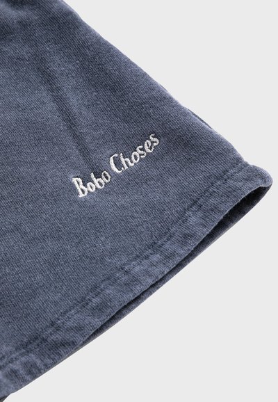Coin de tissu bleu avec texte brodé blanc "Bobo Choses" près de l'ourlet, montrant les détails de la texture et des coutures.