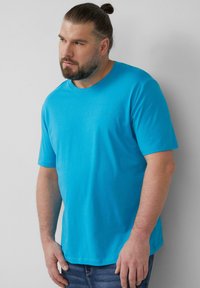 Turkusowa koszulka z krótkim rękawem, wykonana z miękkiego materiału, z okrągłym dekoltem i subtelnym logo na klatce piersiowej. Połączona z dżinsowymi spodniami.