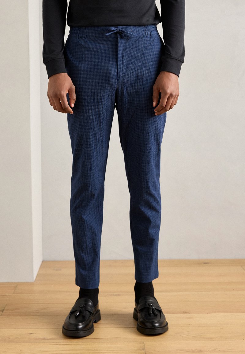 Pantalon en navy texturé avec taille à cordon, coupe fuselée, associé à des mocassins foncés et des chaussettes noires, sur un fond clair.