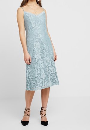 Kobieta ubrana w niebieską, koronkową, midi sukienkę bez rękawów z cienkimi ramiączkami oraz czarne, paskowe wysokie szpilki na jednolitym tle.