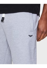 Grijze sweatpants met een zachte textuur, voorzien van zijzakken en een klein zwart geborduurd logo nabij de heup. Witte trekkoord in de taille.