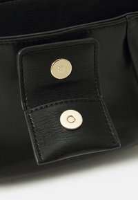 Detail černé kožené kabelky s hladkým povrchem, dvěma zlatými patentkami a úhledným prošitím kolem okrajů.