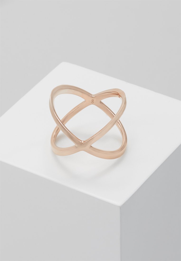 Roze gouden geometrische open ring met kruisende banden, gepresenteerd op een wit kubusvormig voetstuk tegen een grijze achtergrond.