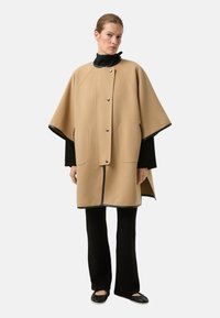 Beige kappecoat lavet af tekstureret stof, med trykknapper, to store forlommer og sort kantdetalje. Båret over sort tøj.