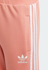 Los pantalones de chándal Adidas en un suave tono rosa claro presentan franjas laterales blancas y un pequeño logo en el bolsillo, exhibiendo una tela suave y un diseño casual.