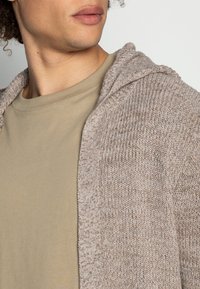 Lichtbruine gebreide cardigan met capuchon, textuurpatroon, gedragen over een beige shirt. De cardigan heeft een open voorzijde.