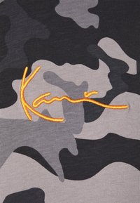 Tkanina s maskáčovým vzorem v šedé, černé a bílé barvě, s oranžovým vyšitým textem "Kani" v vlnitém písmu napříč středem.