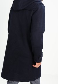 Cappotto in lana blu navy con cappuccio, dotato di chiusura con bottoni, due tasche laterali e una finitura leggermente strutturata. Maniche lunghe con polsini abbottonati.