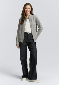 Grijze zip-up jack met een textuurafwerking, gedragen over een wit shirt en donkerblauwe wijde jeans. De outfit heeft lichte schoenen.