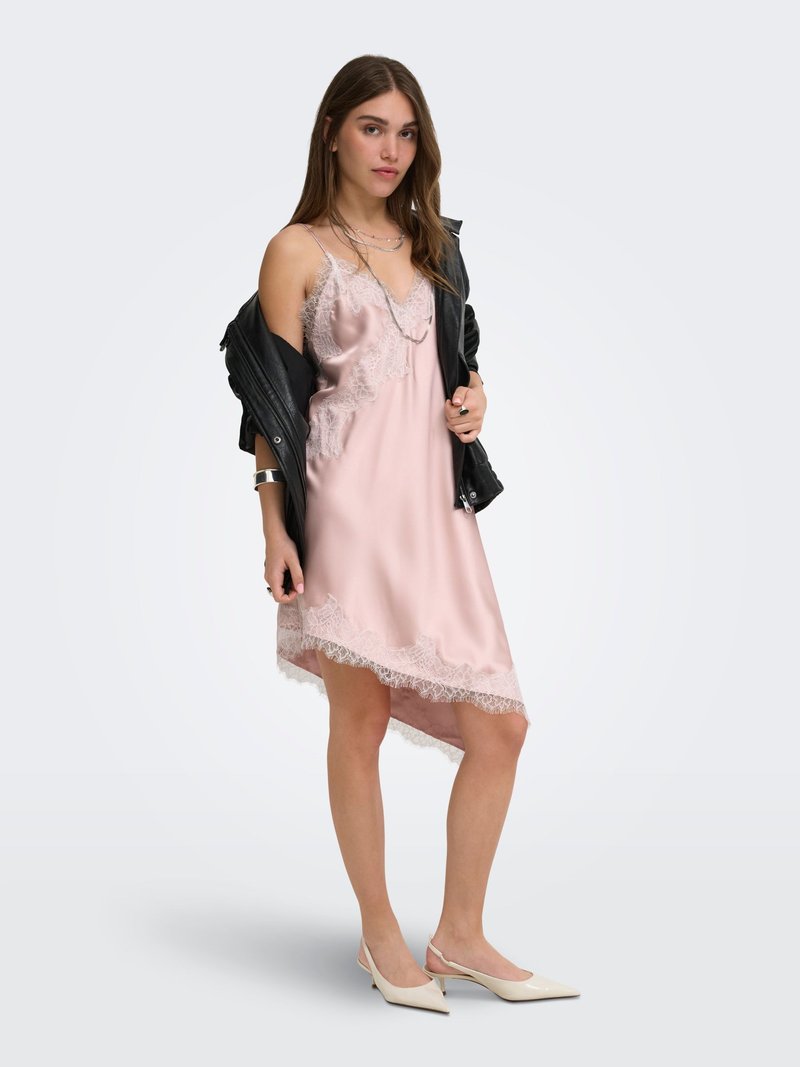 Jeune femme portant une robe nuisette rose clair bordée de dentelle, des talons beige à bride arrière et une veste en cuir noire posée sur ses épaules.
