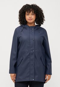 A woman with curly hair wears a dark blue raincoat with snap buttons and a hood, posing against a plain background.