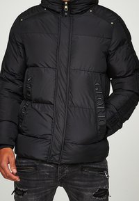 Černá péřová bunda se zipem vpředu a nápisem "Glorious" vyšitým na kapsách, nošená s potrhanými černými džínami s ozdobnými zipy.