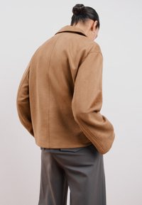 Ruskea keinonahkainen takki, jossa on lovellinen kaulus, ylisuuria hihoja ja lyhyt malli, yhdistettynä harmaisiin housuihin.