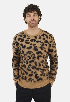 Homme aux cheveux bouclés portant un pull à imprimé léopard marron et noir, les mains dans les poches de son pantalon noir, se tenant devant un fond uni.