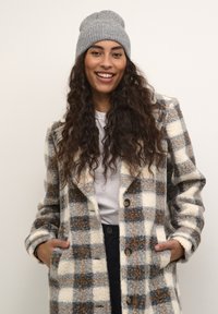 Manteau à carreaux en crème, gris et marron, fabriqué en tissu texturé ; il présente de grands boutons et des poches latérales. Associé à un bonnet en tricot gris.