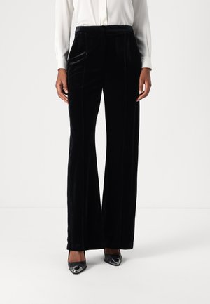 Pantaloni a gamba larga in velluto nero con pieghe frontali, abbinati a una camicia bianca con bottoni. La persona indossa tacchi a punta neri.