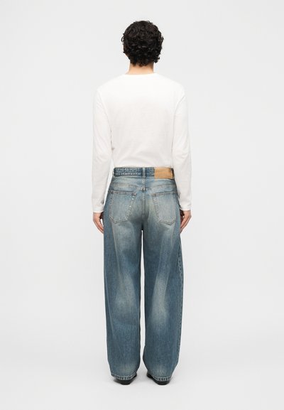 Άτομο με σγουρά σκουρόχρωμα μαλλιά, φορώντας λευκό πουκάμισο με μακριά μανίκια και χαλαρά ανοιχτό μπλε τζιν, στέκεται με την πλάτη γυρισμένη σε απλό φόντο.