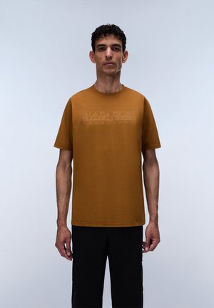 Homme aux cheveux courts et bouclés portant un t-shirt marron moutarde à manches courtes avec un texte imprimé discret, se tenant devant un fond gris clair uni.