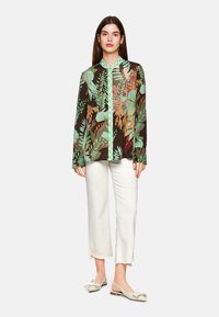 Long-sleeve shirt featuring a multicolor tropical leaf pattern on a dark background, with a green collar and button closure. Paired with white pants.