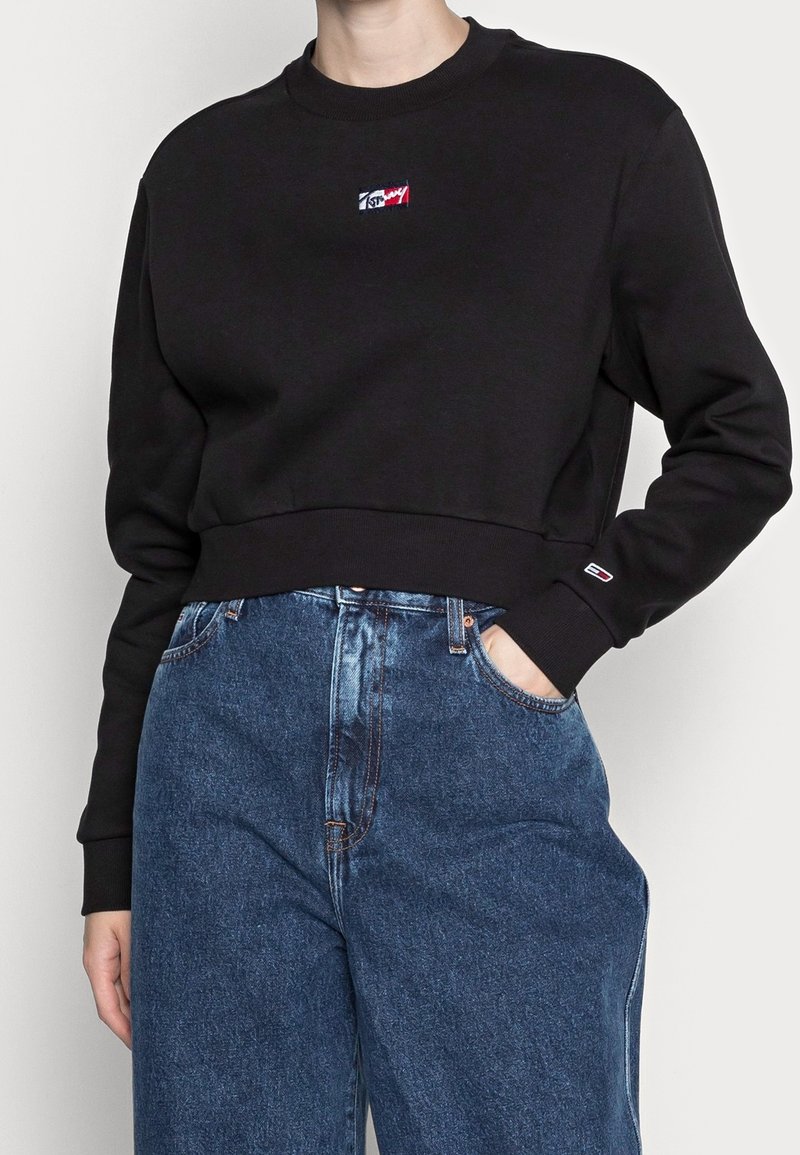 Persona vestida con un sudadera corta negra con un pequeño logo en el pecho y en el puño, acompañada de unos vaqueros de mezclilla azul de alta cintura, con la mano en el bolsillo.