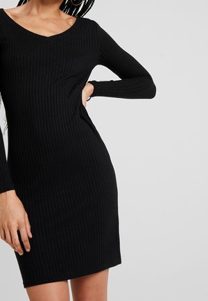 Vrouw draagt een zwarte, geribbelde, lange mouwen, knie-lange jurk met een V-hals, staand met één hand op haar heup.