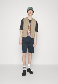 Gilet beige sur un t-shirt noir avec un graphique coloré, associé à un short marine. Les accessoires comprennent une casquette verte et des baskets noires.