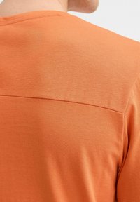 Majica kratkih rukava s okruglim izrezom u vibrantnoj narančastoj boji, koja se ističe teksturiranim materijalom i glatkim kontrastnim panelom duž stražnjeg ramena.