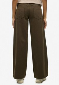 Vrouw die wijde donkerbruine broek en witte sneakers draagt, gezien van achteren tegen een effen achtergrond.