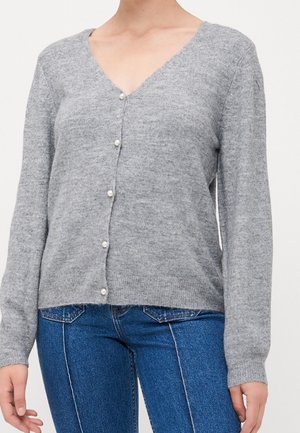Femme portant un cardigan gris clair à col en V avec des boutons en perles et un jean bleu, debout devant un fond uni.