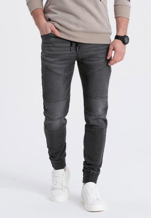 Man wearing black ribbed biker jeans, white sneakers, beige sweatshirt, and black wristwatch, standing with one hand in pocket.