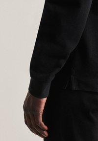 Czarna bluza z teksturowanej tkaniny, z prążkowanymi mankietami i bocznym rozcięciem. Ręka spoczywająca obok odzieży, prezentująca odcień i strukturę skóry.