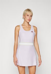 Mulher loira vestindo um vestido sem mangas de tom roxo claro com um cinto branco, de pé contra um fundo branco liso, olhando para a frente.