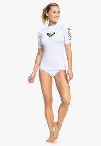 Bela majica s kratkimi rokavi za surfanje z black heart logotipom na sredini in "Roxy" napisom na rokavu; gladek, raztegljiv material.