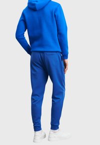 Person som bär en klarblå hoodie och matchande joggerbyxor, står med en hand på höften, vita sneakers synliga, enkel ljus bakgrund.