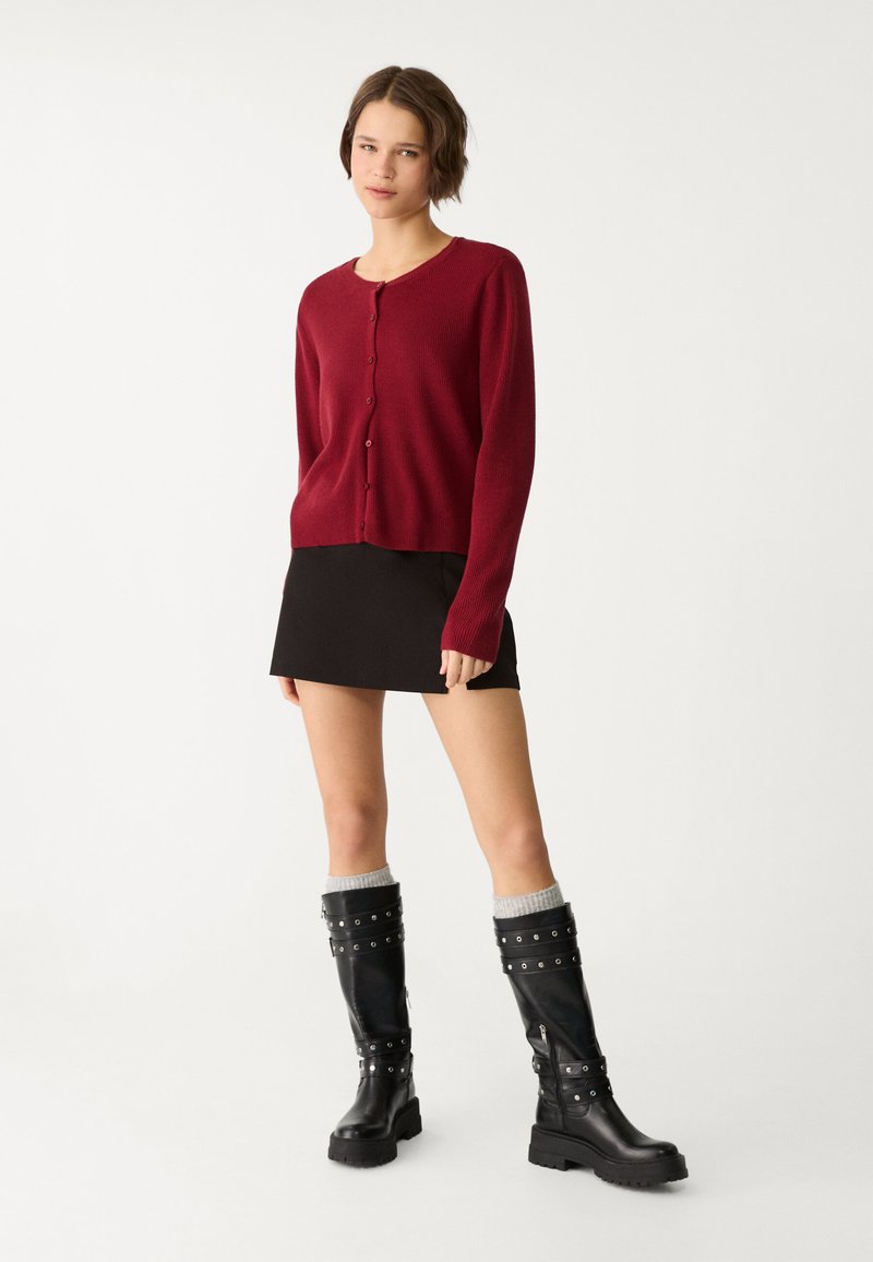 Roter gerippter Cardigan mit Rundhalsausschnitt und Knopfverschluss, kombiniert mit einem schwarzen Minirock und knielangen schwarzen Lederstiefeln mit silbernen Akzenten.