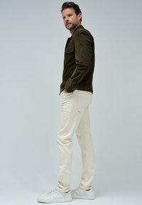 Homme se tenant de côté portant une veste en daim marron foncé, un jean écru et des baskets blanches, sur un fond léger uni.