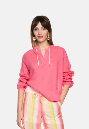 Frau trägt eine leuchtend pinke langärmelige Bluse mit Kordeln und pastell gestreifte Hose, steht mit einer Hand in der Tasche vor weißem Hintergrund.