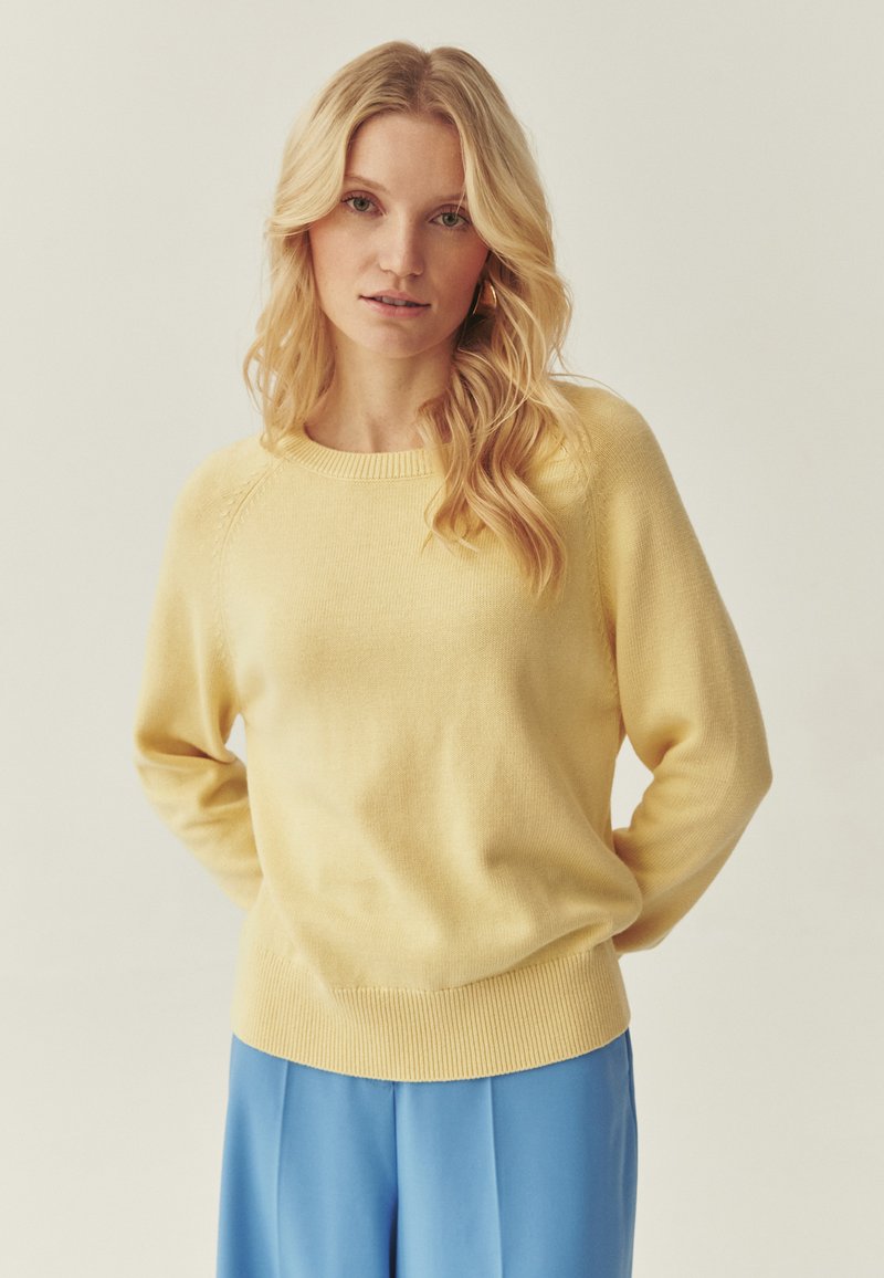 Pull jaune en tricot avec encolure ronde et ourlet côtelé, associé à un pantalon bleu. Texture douce avec une coupe décontractée et des détails subtils.